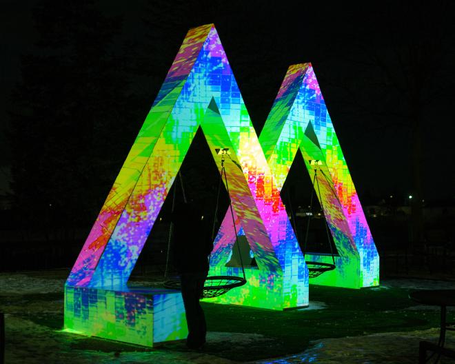 A sculpture in the shape of an &lsquo;M&rsquo; with a variety of colors, patterns, and shapes being project onto it. It&rsquo;s striking against the night sky.