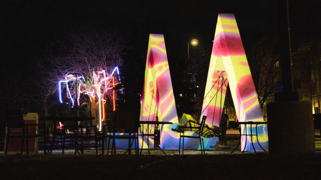 A sculpture in the shape of an &lsquo;M&rsquo; with a variety of colors, patterns, and shapes being project onto it. It&rsquo;s striking against the night sky.