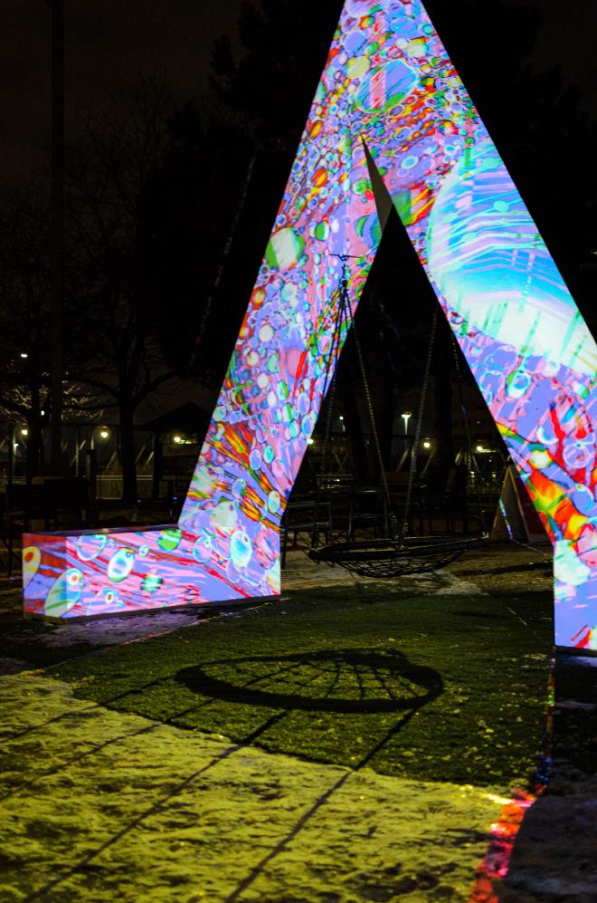 A sculpture in the shape of an &lsquo;M&rsquo; with a variety of colors, patterns, and shapes being project onto it. It&rsquo;s striking against the night sky.