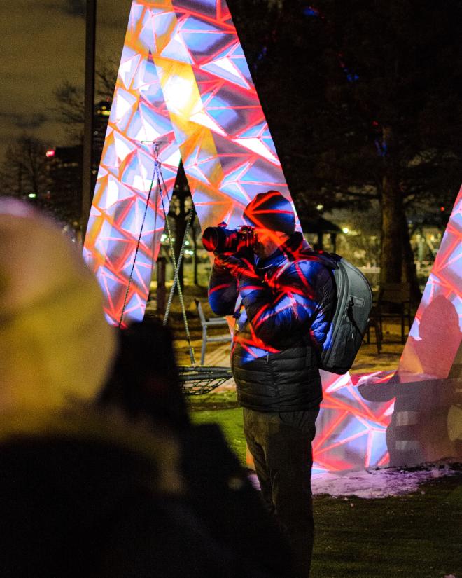 A sculpture in the shape of an &lsquo;M&rsquo; with a variety of colors, patterns, and shapes being project onto it. It&rsquo;s striking against the night sky.