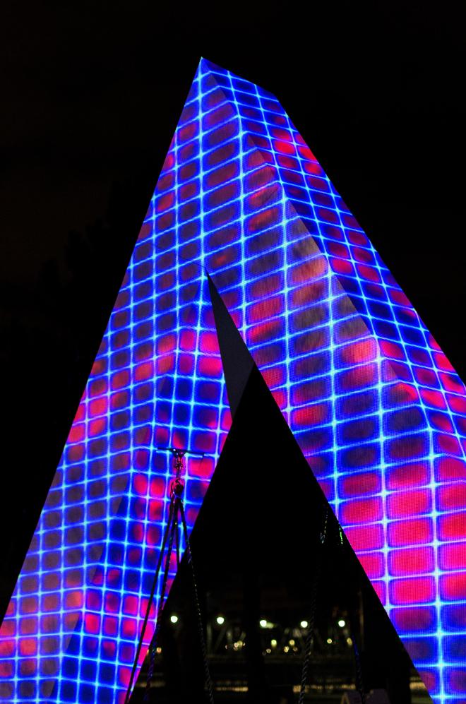 A sculpture in the shape of an &lsquo;M&rsquo; with a variety of colors, patterns, and shapes being project onto it. It&rsquo;s striking against the night sky.