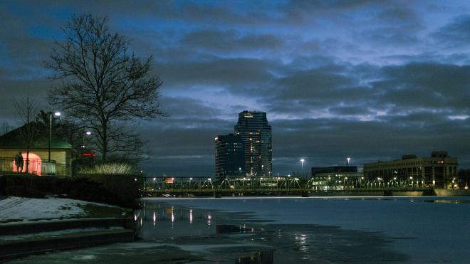 a tall glass and steel building in the distance, with a wide urban river frozen over with snow on top in the foreground. The vibes are cold, blue, and dark.