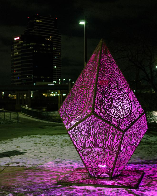 an angular, diamond-shaped sculpture with symbols of the Native American community throughout. There is a glowing light that changes color lighting the sculpture from within.