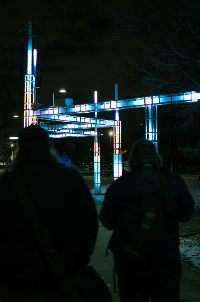 an angular structure that makes its way along and over a sidewalk, each piece and pillar contains LED lights that change color